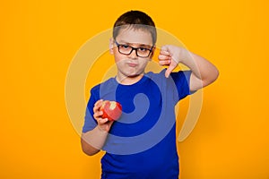 Portrait of a boy in glasses and a blue T-shirt stands on a yellow background and holds a tasteless apple in his hand