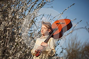 Portrait boy with guitar standing near blooming flowers in summer day.