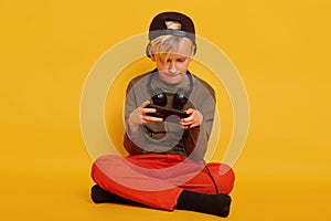 Portrait of boy, playing online video games via his modern smartphone isolated over yellow background, looks concetrated on his
