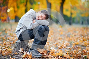 Portrait of boy sitting and dreaming on a stump in autumn city park. Posing on trees with yellow leaves. Bright sunlight and