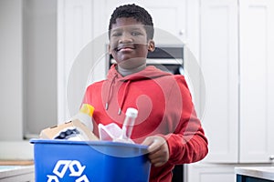 Portrait Of Boy Standing In Kitchen At Home Carrying Recycling Bin