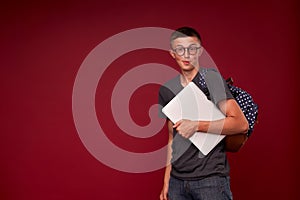 Portrait of a boy student with a backpack and laptop in his hands smiling on a red background. funny positive teenager is a high