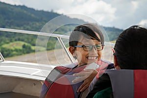 A portrait of boy wearing glasses and life jacket smiling to other child on a boat. Cruising outdoor