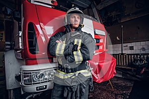 Young fireman wearing protective uniform standing next to a fire engine in a garage of a fire department