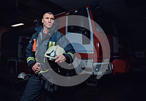 Young fireman wearing protective uniform standing next to a fire engine in a garage of a fire department