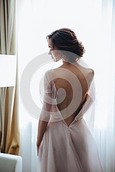 Portrait of the bride with open dress from the back on the window background. Beautiful neckline on the dress with sexy