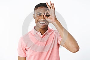 Portrait of bright and cheerful young dark-skinned guy in pink polo shirt showing okay or zero gesture over eye looking