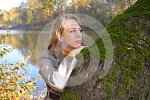 Portrait of a brooding woman leaning against an old suede tree against the background of a lake. Autumn Park