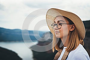 Portrait of brooding young girl with blonde hair in straw summer hat and hipster glasses on backdrop river natura mountain land