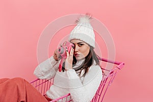 Portrait of brown eyed woman in white hat and sweater taking picture on instax while sitting in supermarket trolley on