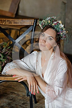 Portrait of a brunette bride with a wedding hairstyle and a flower wreath, in a white transparent negligee. Morning of the bride