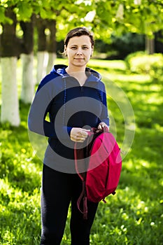 Portrait of brunette girl in sport suit with backpack in green summer park. girl is looking at the frame in bright sunny day