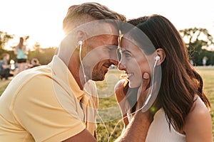 Portrait of brunette middle-aged couple man and woman kissing and listening to music together with earphones in summer park