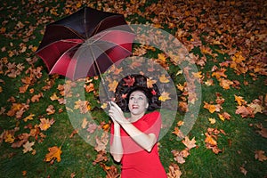 Portrait of a brunette woman with long hair. She lies on green grass in a red dress and maple leaves in her hair holds