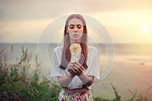 Portrait of a brunette young woman with a big dandelion on a background of warm sunset. summer, outdoors.