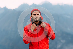 Portrait of a brutal bearded man in a red jacket and hat among the mountains. Portrait of a traveler