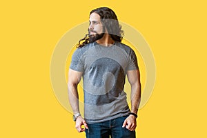 Portrait of brutal handsome bearded young man model with long curly hair in grey tshirt standing and looking away with serious