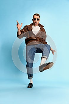 Portrait of brutal, handsome man in jeans, leather jacket and sunglasses posing, dancing over blue studio background