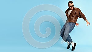 Portrait of brutal, handsome man in jeans, leather jacket and sunglasses posing, dancing over blue studio background