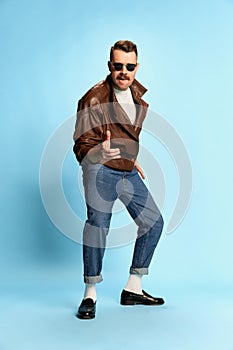 Portrait of brutal, handsome man in jeans, leather jacket and sunglasses posing, dancing over blue studio background