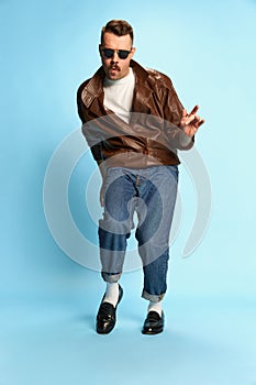 Portrait of brutal, handsome man in jeans, leather jacket and sunglasses posing, dancing over blue studio background