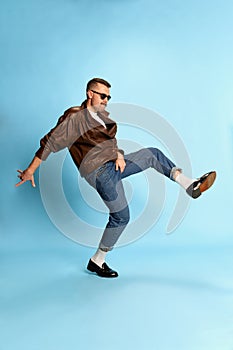 Portrait of brutal, handsome man in jeans, leather jacket and sunglasses posing, dancing over blue studio background