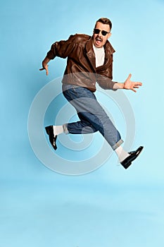 Portrait of brutal, handsome man in jeans, leather jacket and sunglasses posing, jumping over blue studio background