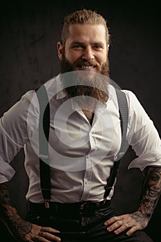 Portrait of a brutal man with a beard and long hair he looks at the camera and smiles dressed in a shirt and jeans with suspenders