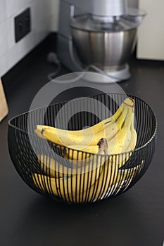 A portrait of a bunch of bananas lying in a decorative black metal bowl on a black kitchen counter. The healthy energizing food is