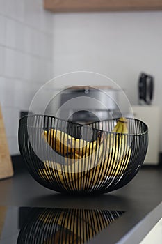 A portrait of a bunch of bananas lying in a decorative black metal bowl on a black kitchen counter. The healthy energizing food is