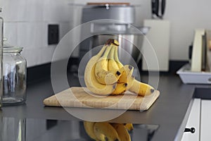A portrait of a bunch of yellow bananas lying on a wooden cutting board on a black kitchen countertop. The delicious energizing