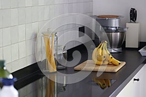 A portrait of a bunch of yellow bananas lying on a wooden cutting board on a black kitchen countertop. The delicious energizing