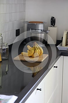 A portrait of a bunch of yellow bananas lying on a wooden cutting board on a black kitchen countertop. The delicious energizing