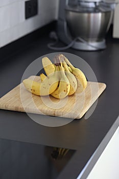 A portrait of a bunch of yellow bananas lying on a wooden cutting board on a black kitchen countertop. The delicious energizing