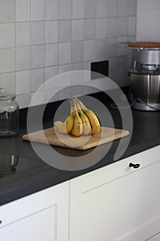 A portrait of a bunch of yellow bananas lying on a wooden cutting board on a black kitchen countertop. The delicious energizing