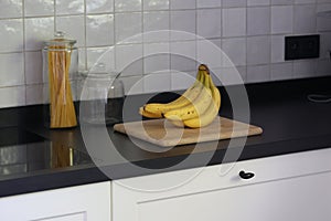 A portrait of a bunch of yellow bananas lying on a wooden cutting board on a black kitchen countertop. The delicious energizing