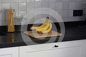 A portrait of a bunch of yellow bananas lying on a wooden cutting board on a black kitchen countertop. The delicious energizing
