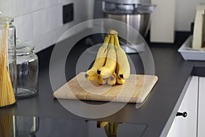 A portrait of a bunch of yellow bananas lying on a wooden cutting board on a black kitchen countertop. The delicious energizing