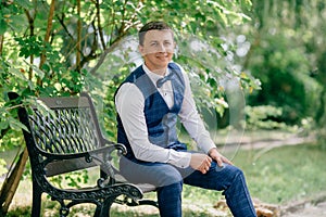 Portrait of business man in suit sitting in park on bench and smiling. The guy is getting ready for the wedding day