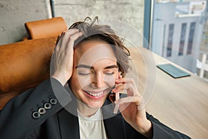 Portrait of business woman in suit, sitting in her office and answering a phone call with pleased smile, having a