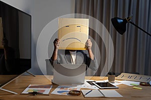 Portrait of businessman covering his face with paper mask with sad face drawn on it, bad negative