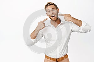 Portrait of businessman pointing at his white teeth, perfect whitened smile, dental clinic advertisement, standing over