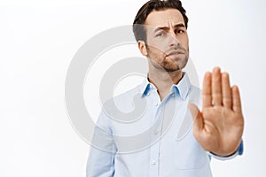 Portrait of businessman showing stop sign, disapprove smth, extends his palm in rejection, stands over white studio