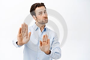 Portrait of businessman showing stop sign, disapprove smth, extends his palm in rejection, stands over white studio