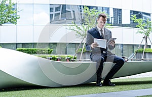 Portrait of businessman sitting on a bench and holding tablet and file at business center. Concept of work life balance