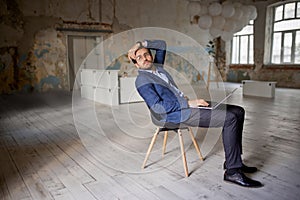 Portrait of businessman sitting on chair in empty room and working on laptop. Making professional analytics and