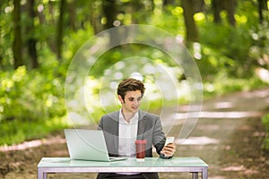 Portrait of Businessman sitting at the office desk with laptop computer and cup of coffee talking mobile phone in green forest par