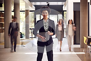 Portrait Of Businesswoman With Digital Tablet Standing In Lobby Of Busy Modern Office Building