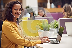 Portrait Of Businesswoman Wearing Phone Headset Talking To Caller In Busy Customer Services Centre