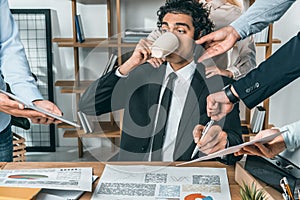 portrait of busy businessman drinking coffee and sitting at workplace while colleagues helping with work